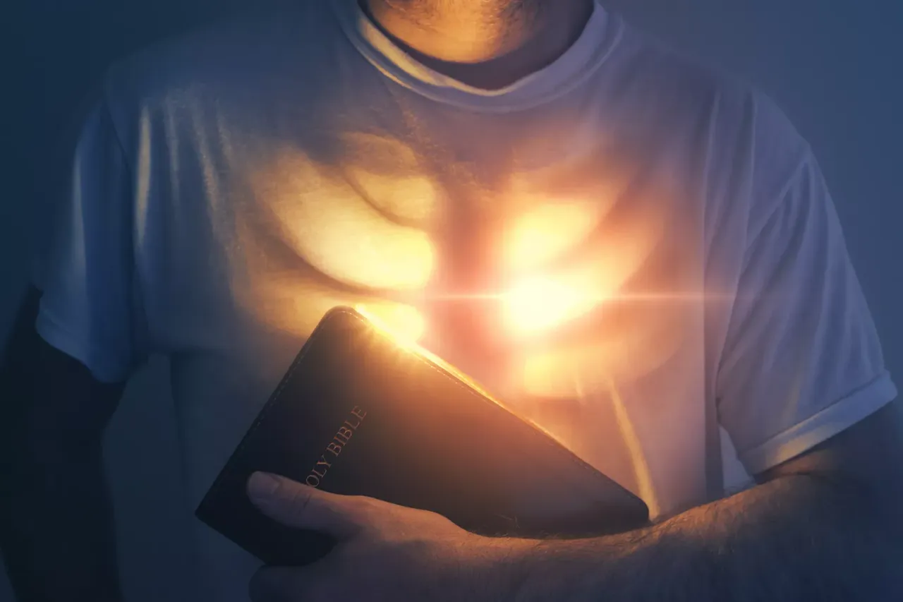 Eine Person hu00e4lt eine Bibel vor der Brust, welche warm leuchtet wie auch das Herz der Person.